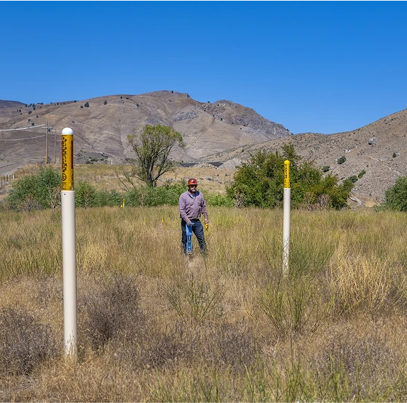 Man standing in a grassy field between pipeline markers with mountains in the background