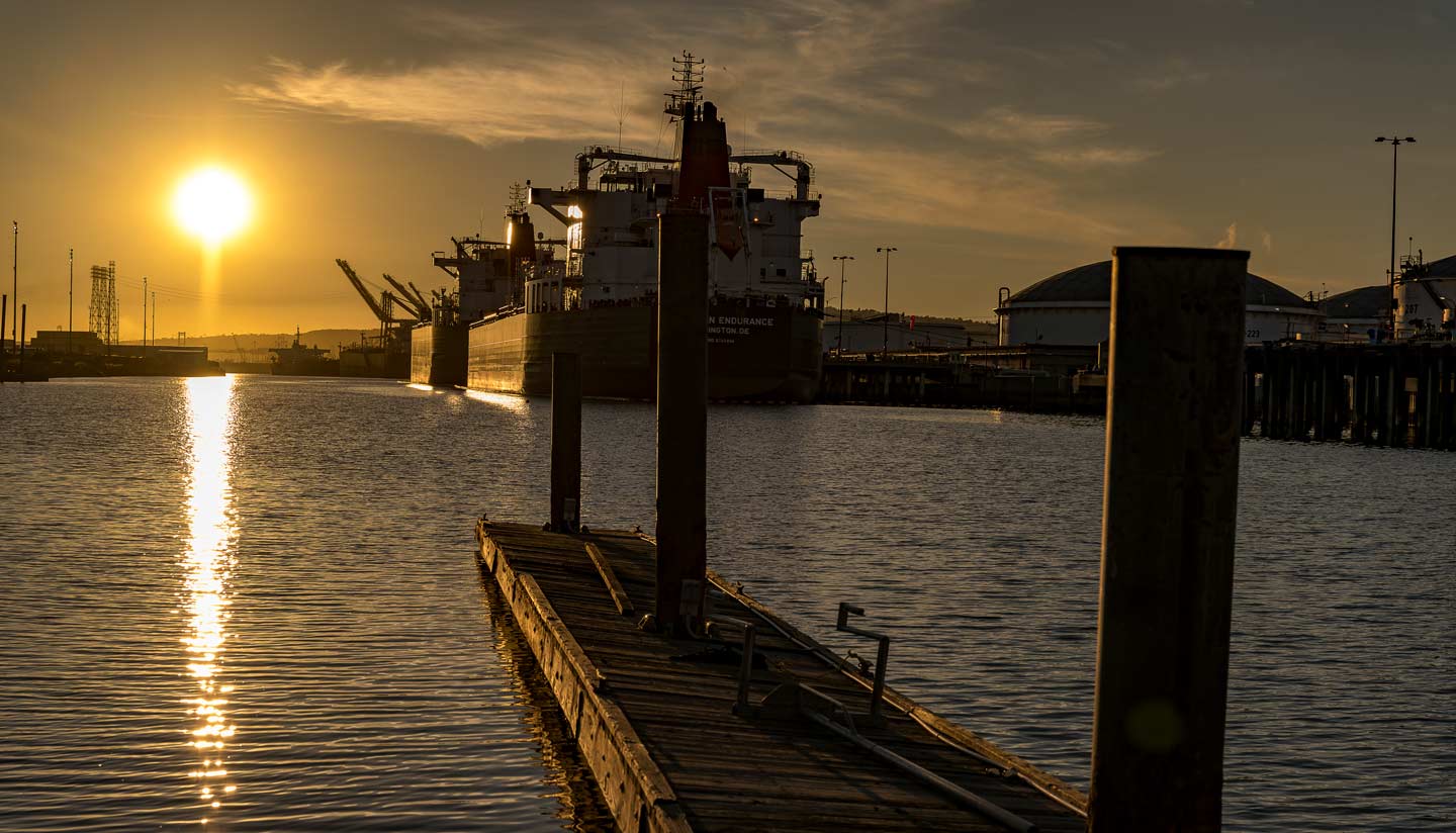 Large tanker ship docked at an industrial port during sunset, with the sun reflecting on the water and a wooden pier in the foreground