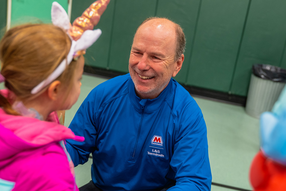 An adult leans down and smiles at a child indoors after helping with a coat.