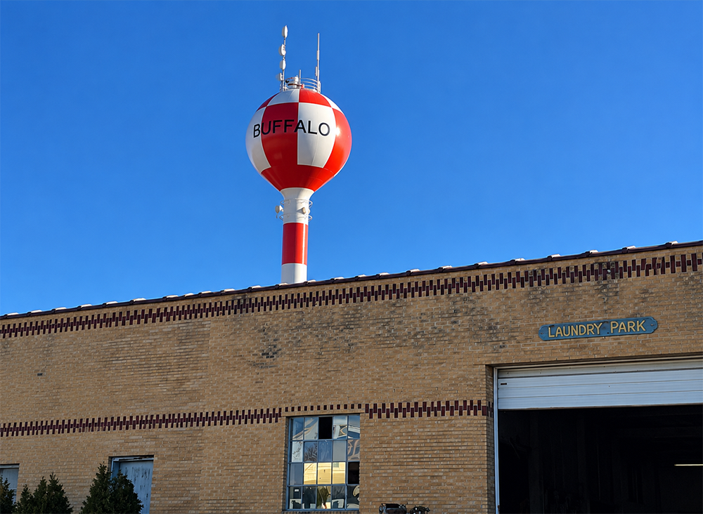 A red-and-white water tower with "BUFFALO" written across it rises above a brick building against a clear blue sky.