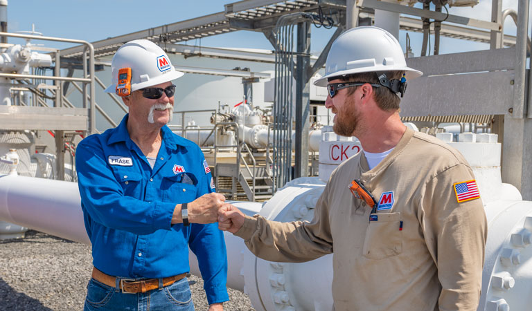 Two workers wearing hard hats and safety gear bump fists at an industrial facility with large pipes and metal structures in the background.