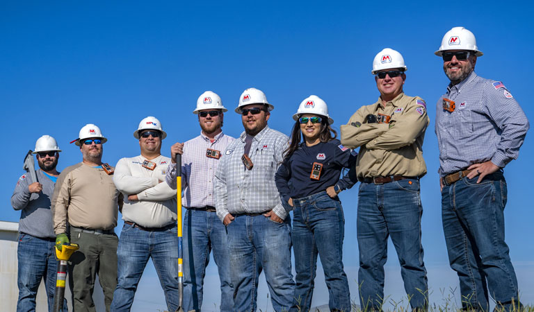 Eight utility workers wearing hard hats and safety gear stand together outdoors under a clear blue sky, holding equipment and tools.