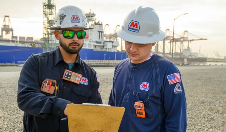 Two MPL dock workers reviewing paperwork on a clipboard with a large tanker in the background