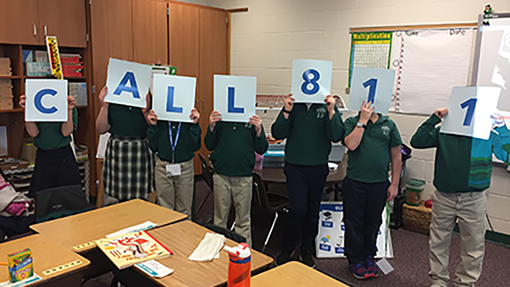Seven children in a classroom stand in a row holding up large signs that spell out "CALL 811," with their faces mostly hidden behind the signs.