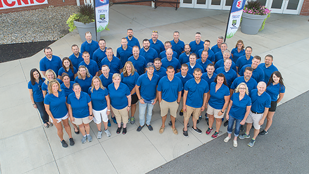 A large group of people wearing matching blue shirts stands together on a paved area outside a building with banners and large flower pots.