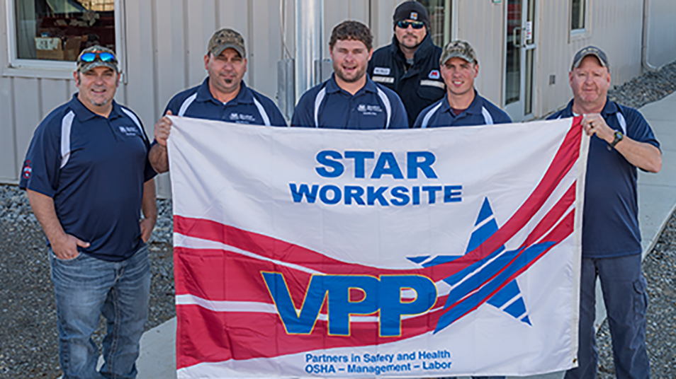 Six people in matching navy blue shirts stand outside a metal building, holding a banner that reads "STAR WORKSITE VPP Partners in Safety and Health OSHA &ndash; Management &ndash; Labor" with a red and blue star design.