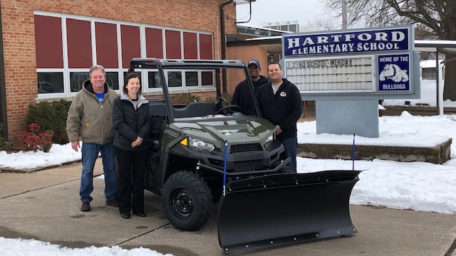 Four adults stand next to a green utility vehicle with a snow plow attachment in front of Hartford Elementary School, with snow on the ground and a school sign visible in the background.