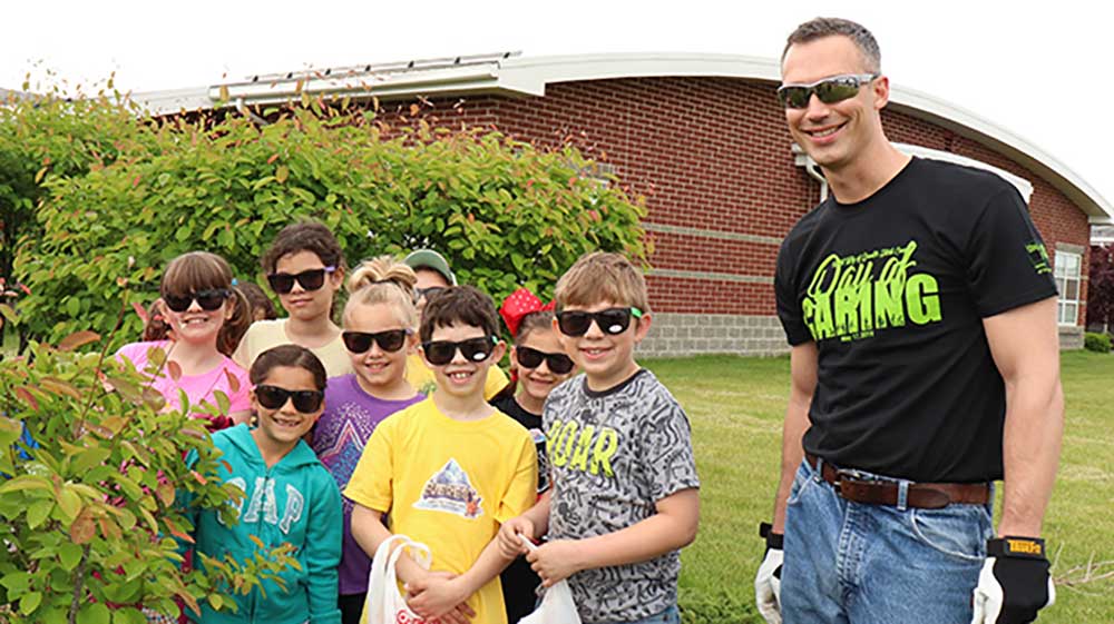 A group of children stands outdoors near green bushes with an adult wearing gloves and a "Day of Caring" t-shirt, in front of a brick building with a curved roof.