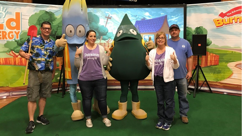 Five adults stand on a green carpet in front of a colorful backdrop with cartoon scenery, posing with two large costumed mascots-one shaped like a blue flame and the other a green drop-while one person holds a red shovel and two others give thumbs up.