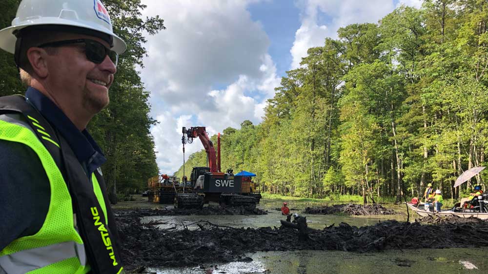 A construction worker in a safety vest and hard hat observes a swampy worksite with heavy machinery and other workers in the background, surrounded by dense green trees under a partly cloudy sky.