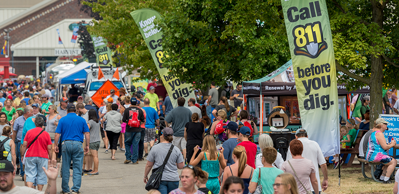 A large crowd walks through an outdoor fair with informational booths and tall green banners that read "Call 811 before you dig," surrounded by trees and various displays.