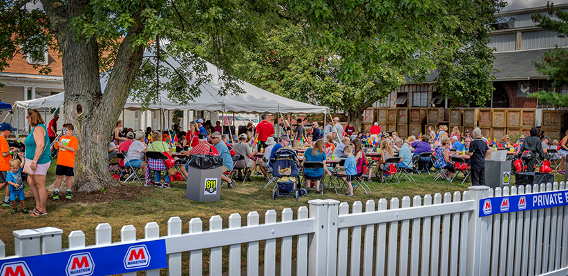 Groups of people sit at tables under a large white tent and nearby trees at an outdoor event, with children and adults socializing and an "811" sign visible on a trash bin in the foreground.
