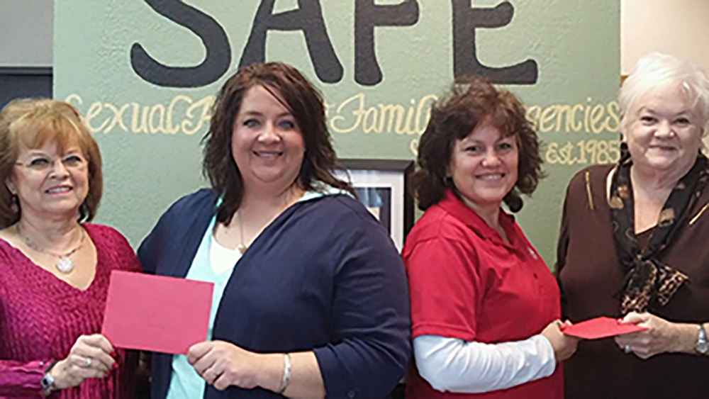 Four women stand indoors holding red envelopes in front of a wall with the word "SAFE" and additional text about sexual assault and family emergencies.