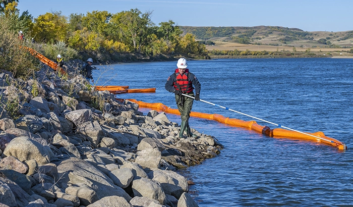 An MPL emergency official using a long poll to pull a long row of orange inflatable tubbing across water at a rocky shoreline