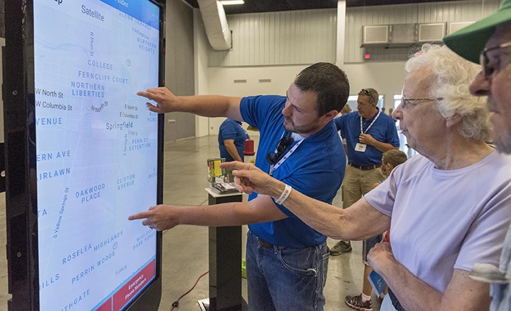 An MPL employee in a blue shirt assisting an elderly woman as they point to pipeline locations on a large interactive map display at an indoor event