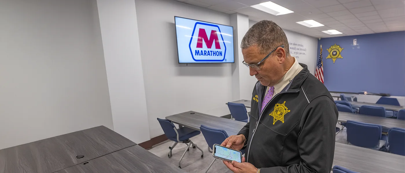 A sheriff standing in a conference room with the Marathon Petroleum logo displayed on a TV, he is looking at down at emergency response details for pipeline incidents