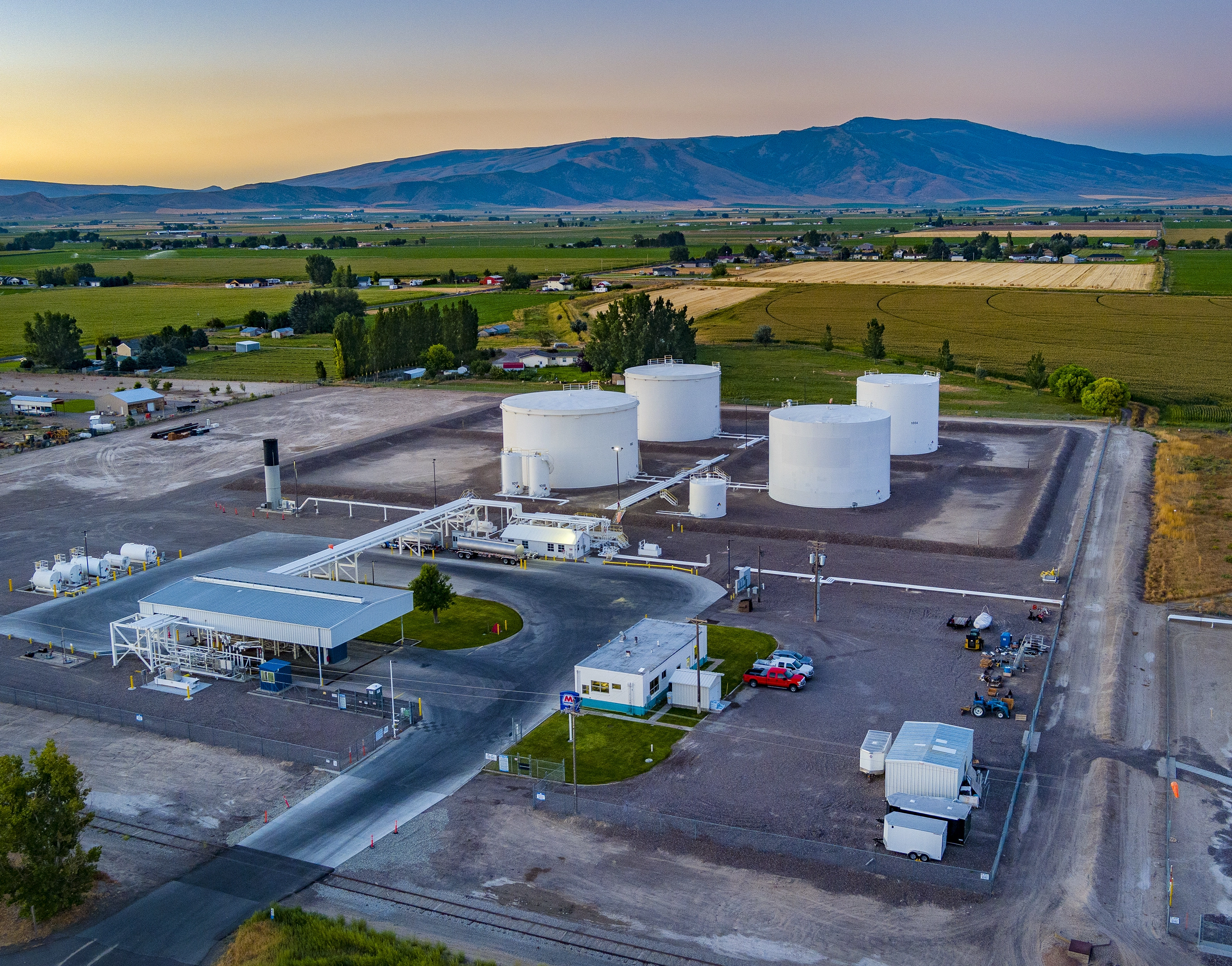 An aerial view of a rural fuel storage facility with several large white storage tanks, surrounded by farmland and distant mountains at sunset