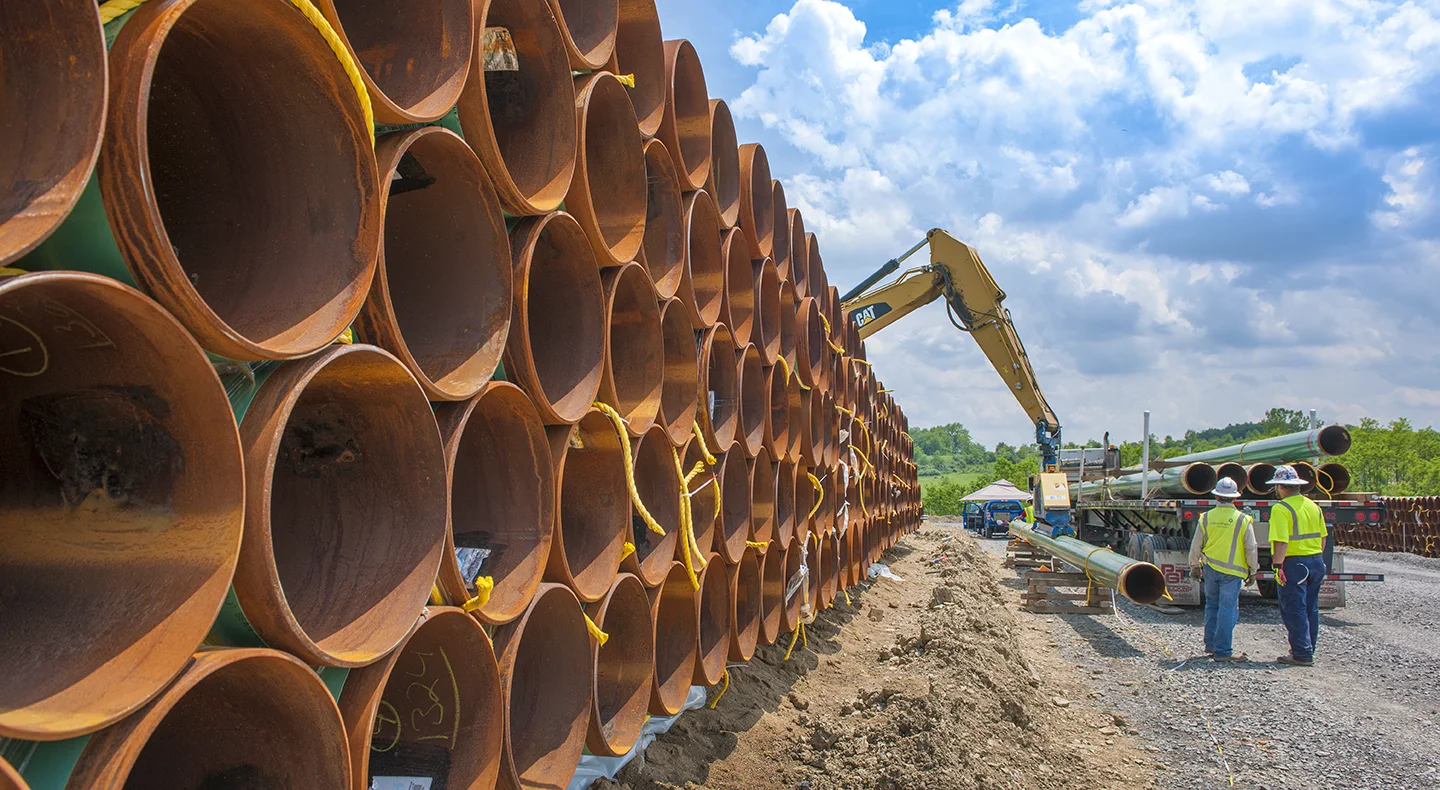 Huge brass pipes stacked next to a large semi-truck with silver pipes and construction workers at a pipeline construction site