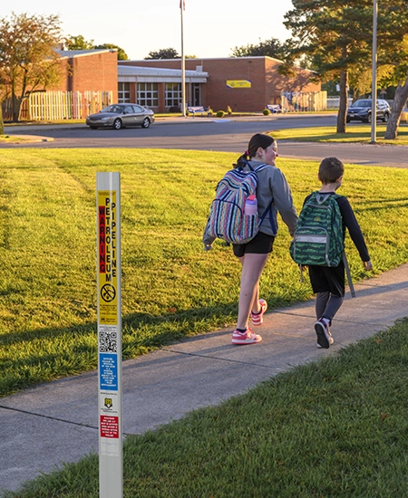 Two children with backpacks walking on a sidewalk past a petroleum pipeline marker near a school building