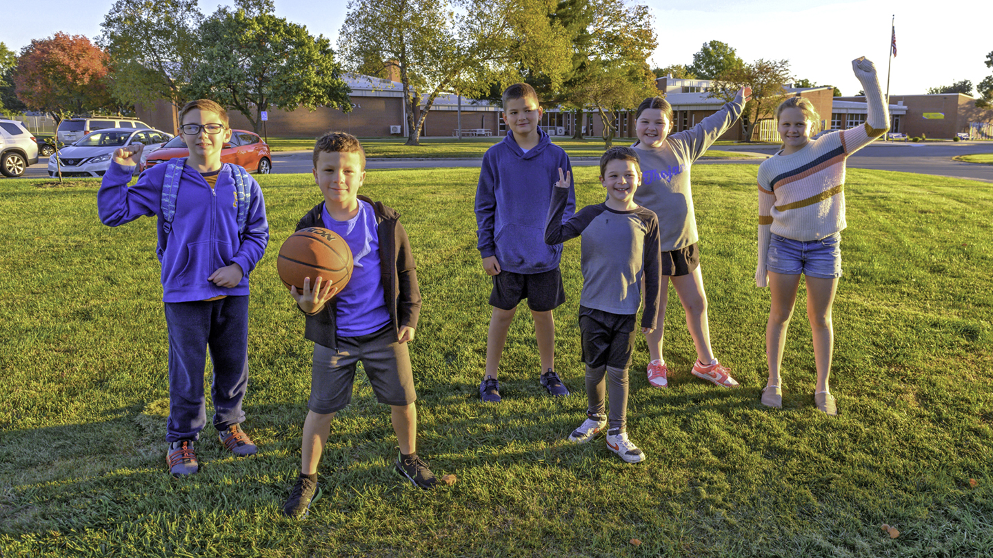 Group of six children posing and playing with a basketball on a grassy field in front of a school building on a sunny day