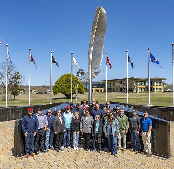 A group of people from MPL and the Osage nation pose for a photo in front of a large feather sculpture surrounded by flagpoles, with a modern building and trees in the background on a clear, sunny day