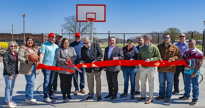 A group of people stands in front of an outdoor basketball hoop holding a large red ribbon getting ready to cut it