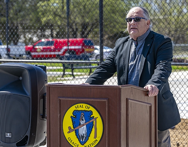 An Osage leader stands at a podium displaying the Seal of Osage Nation, speaking outdoors near a large speaker, with a chain-link fence and a red emergency vehicle in the background