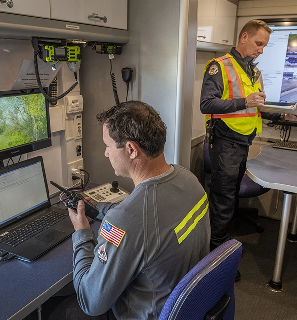 Two emergency response personnel operate communication and monitoring equipment inside a mobile command center
