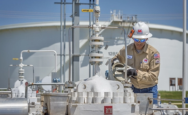 A man in a hard hat and protective gear maintaining equipment on a pipeline with a tanker and clear sky behind him