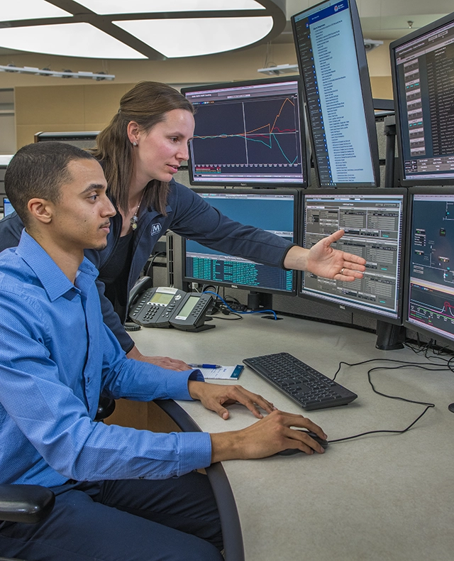 Two MPL employees working at a pipeline control center desk with multiple computer monitors displaying data and graphs.