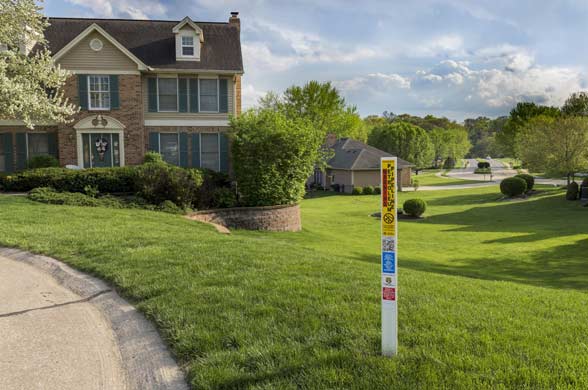 Residential neighborhood with a two-story brick house, green lawn, and a utility marker sign by the roadside marking where a pipeline is