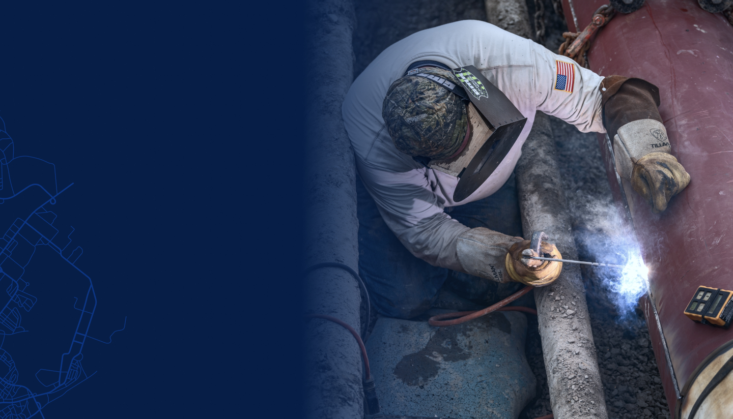 A worker in protective gear sitting amongst muddy pipes, welding a large pipeline