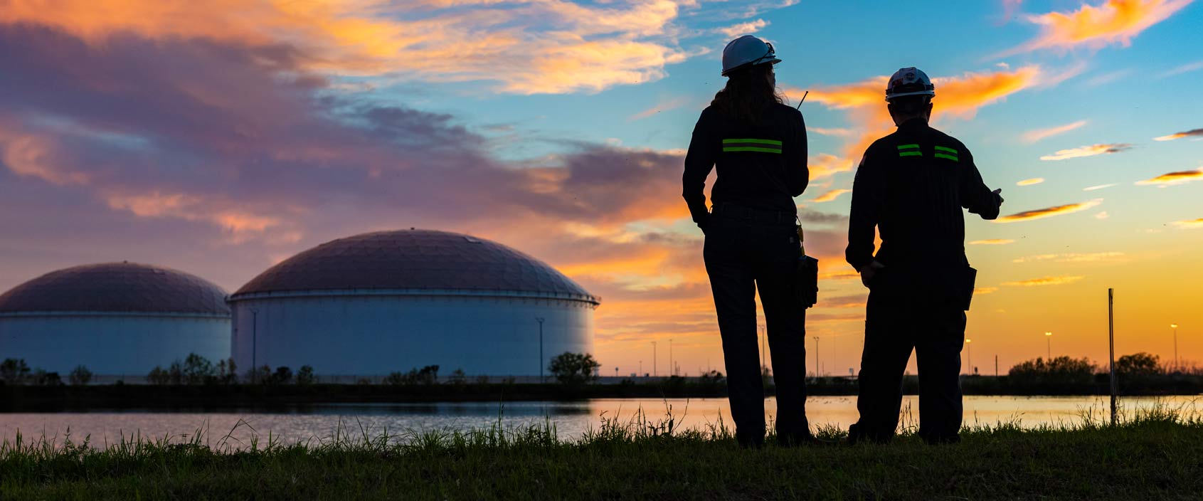 Two workers wearing safety helmets and reflective clothing stand by a waterway at sunset, observing large industrial storage tanks in the distance