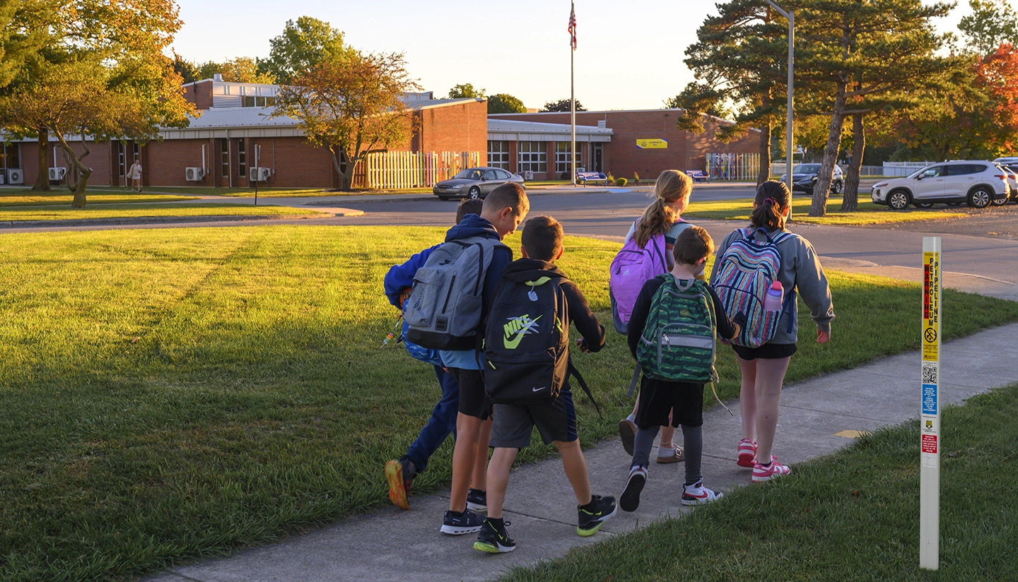 A group of school children with backpacks walk together on a sidewalk toward a brick school building in the morning sunlight