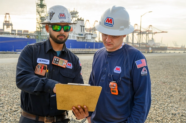 Two MPL dock workers reviewing paperwork on a clipboard with a large tanker in the background